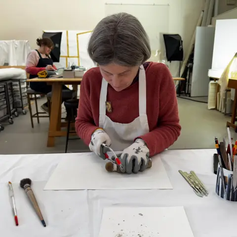 Capital Theatres Helen Creasy has grey bobbed hair. She is wearing a maroon top and white apron. She is also wearing gloves and is cutting the bottle with a small saw.