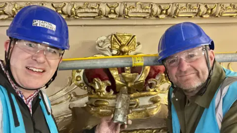 Mike Hume Mike Hume and John Robb wearing blue helmets and safety goggles. They are beside the crown which is painted gold and maroon. There is a scaffolding pole as they are on a raised platform.