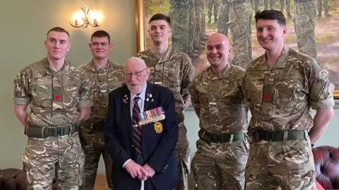 Royal Navy Mr Eskdale seen wearing a blue suit while stood with five recruits all wearing camouflage uniform.