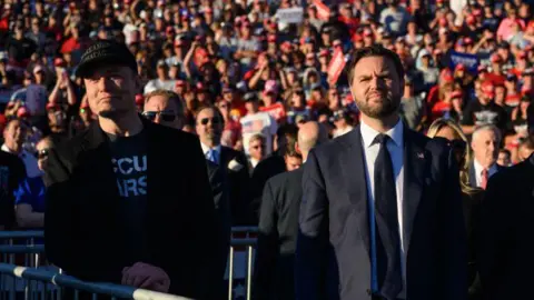 Getty Images Elon Musk and JD Vance look ahead, in front of a crowd, with sun on their faces.