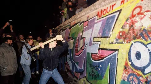 Getty Images A man uses a pickaxe to smash the Berlin wall, surrounded by an enthusiastic crowd.