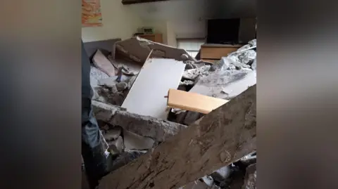 Emyr Owens A damaged bedroom with rubble covering a bed. A large fallen wooden beam can be seen in front of the bed, while two chest of drawers and a window can be seen in the background.