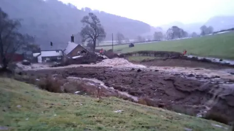 Emyr Owens A brick farmhouse pictured from a distance with tonnes of brown silt covering the yard in front of it. A pile of branches and fallen trees can also be seen by the farmhouse.