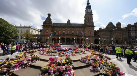 PA Media Floral tributes in a square outside The Atkinson arts centre in central Southport, with large displays left on a circular stone area in the middle of a square in front of an ornate Victorian building in stone, with a clocktower at one end. Peaceful crowds are being marshalled by a handful of police officers