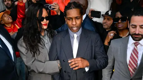 Getty Images A$AP Rocky locks arms with his partner Rihanna outside an LA courthouse as crowds of people and reporters surround them with cameras.