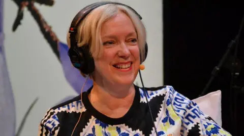 BBC Janice Forsyth wearing a radio headset and microphone. She has short blonde hair and a top with a striking pattern.