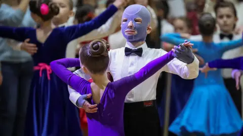 Reuters Roman Oleksiv, wearing a purple burns mask, white shirt and bow tie, dances with girl wearing purple. Many other dancing couples are behind them