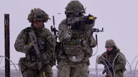 BBC Three soldiers in green camouflage uniforms, holding guns, walking towards a wired fence