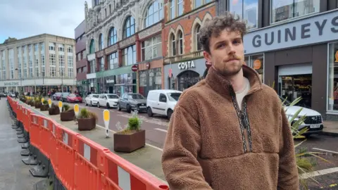 BBC Man wearing a brown teddy material fleece standing in front of an orange temporary barrier along the street. Shops and cars line the other side of the road. 