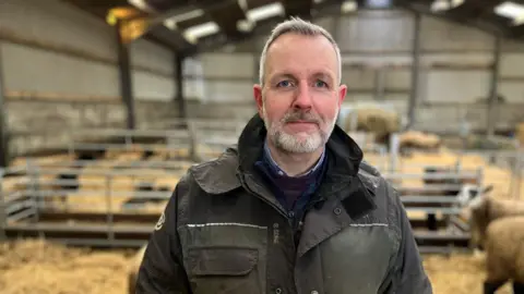 BBC Michael Goldie looking at the camera, wearing a khaki jacket. He is standing in a barn with his surviving lambs and ewes in pens behind him.