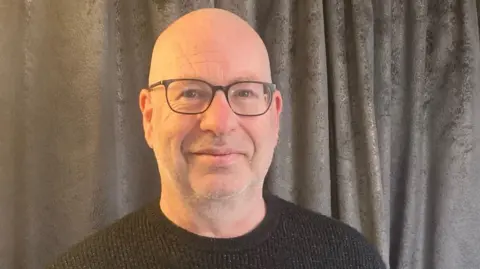 Davy Uprichard Davy Uprichard wearing a grey jumper and glasses looking into the camera with a slight smile. He is standing in front of a grey curtain