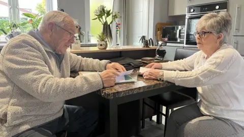 BBC An elderly man and a woman in her 60s sit opposite each other at a marble top kitchen table. They both have folders of paperwork in front of them and they are looking towards each other. Both are wearing glasses. He has white hair and she has grey hair. He is wearing a beige fleece and blue trousers, she is wearing black and white check trousers and a white arran jumper, as well as a large white-faced wristwatch. A window, vase of flowers, microwave and built-in oven are visible behind them.