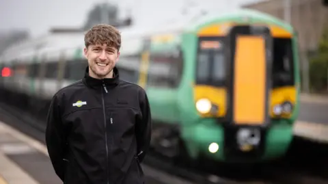 Andrew Gardner/GTR Jake Robinson standing on the platform wearing a black jacket with a green Southern Rail logo on the right. Jake has curly brown hair and a fringe down to his eye brows and is smiling at the camera. There is a green and yellow southern rail train behind him ready to depart from the station