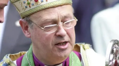 Wales News Service Anthony Pierce wearing a bishop's mitre with a gold trim along the bottom. He is clean-shaven and wearing glasses, and is looking down. The top of his robes and his clerical collar can just be seen in the shot.