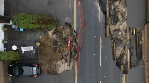 PA Media An aerial view of two sinkholes on a residential road. There is a car on the left which appears to be parked right on the border of one sinkhole.