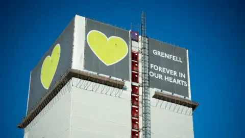 PA Media File image of Grenfell Tower, covered in tarpaulin with a grey and green sign with a heart saying 'Grenfell: Forever in our hearts'