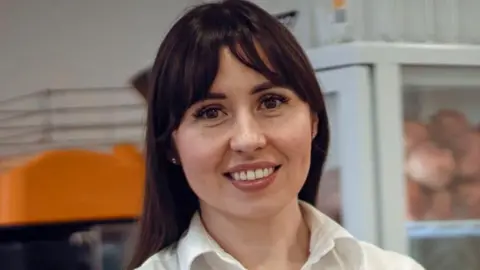 BBC Maryna with brown hair and wearing a white shirt smiles at the camera