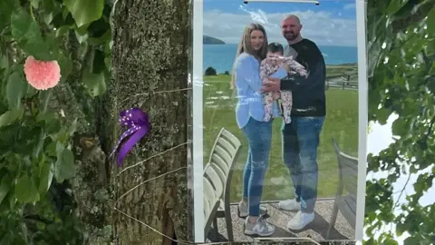 Family handout A photograph of mum Gwen, dad Rob and baby Mabli is tied to a tree. The mum and dad are smiling at the camera holding baby Mabli together in their arms, with the sea in the background. 
