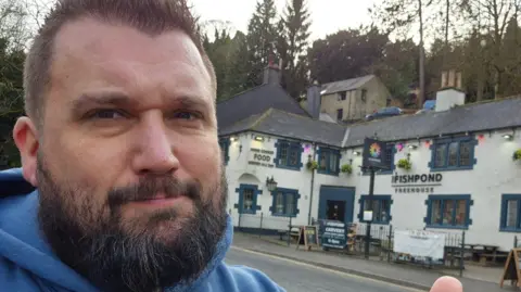The Great British Pub Crawl Dale takes a selfie outside a pub called The Fishpond. It's a white building with navy blue window frames 