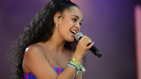 BBC A woman with black curly hair and wearing a pink and blue top singing into a microphone