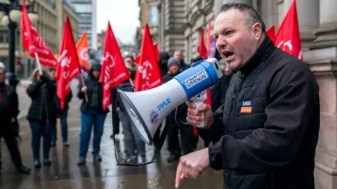 PA Media A man with dark greying hair speaks into a megaphone outside Glasgow City Chambers. He wearing a black jacket with a GMB union badge on it. He is side on to the camera, with protesters holding red flags in the background 