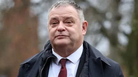 PA Media Mike Amesbury with greying hair, blue raincoat, white shirt and burgundy tie arrives at court.
