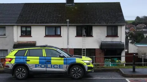 PA Media A yellow and blue police car is parked outside a row of terrace houses. The house on the right has burnt out windows and a ladder leading to the upstairs left window.