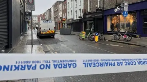 RTE Garda tape in the forefront of the picture in a street in Dublin. A police van is further back in the street as are three parked bicycles and shops.