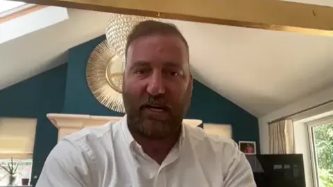 UGC Scott El Paraiso looks at the camera during a June 2024 Zoom meeting with leaseholders. He is tanned, in his early 40s, has short brown hair and a beard and is wearing a white shirt.