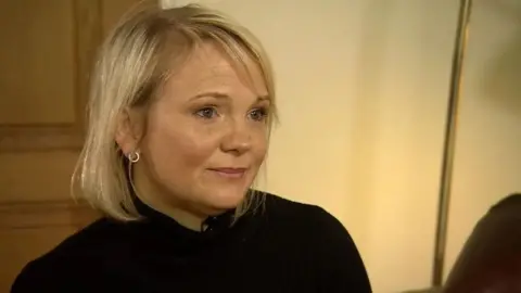 BBC Elizabeth Hudson, a woman in her forties, with blonde hair, circular silver earrings, and a black turtle neck. It is a close-up shot of her head and shoulders, seemingly in her home or a living room.