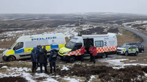 TWSMRT A  police van parked alongside a mountain rescue van. Mountain rescuers can be seen in front of the vans in red jackets with police officers in black standing in a separate group. Moorland can be seen behind the vans which stretches beyond the horizon.