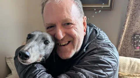 Essex Havard Essex and greyhound Gilly hug in his living room after the news that greyhound racing will be banned in Wales. Essex is wearing a black down jacket while Gilly is grey with a black nose.