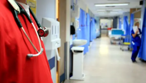 BBC Hospital ward corridor with a medic stood in the foreground - only visible from shoulders to waist, with a red scrub top and a stethoscope. There is a nurse in blue out of focus on the ward, with bays on the ward covered by blue curtains