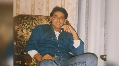 Nottinghamshire Police A photograph of Ethsham Ul-Haq Ghafoor wearing blue denim jeans and a blue shirt sitting on an armchair smiling.