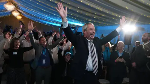 PA Media Reform UK leader Nigel Farage raising his hands in the air and smiling in a organza-clad tent with fairy lights on the ceiling. Lots of supporters are behind him also raising their hands.