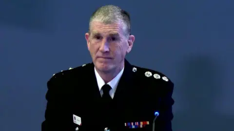 BBC A man is sitting with a navy backdrop behind him. He's wearing a firefighter outfit with medals.