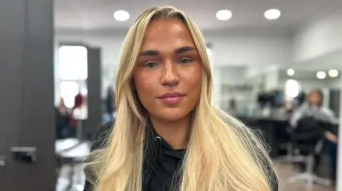 BBC Alex Ritchie pictured in a salon. She has long blonde hair worn loose and wears a black hoodie. Behind her are salon chairs, mirrors, and clients getting their hair cut.