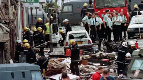 PA Media Several emergency responders are standing in a street in the aftermath of a bomb. Rubble and debris cover the street and road. Cars are damaged with glass blown out