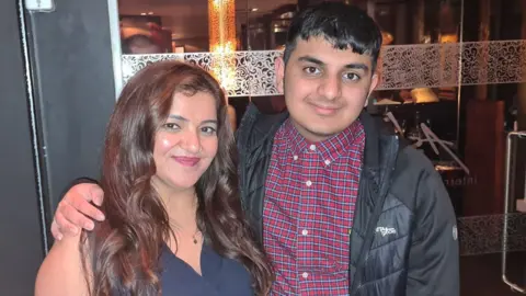 Family Handout Pooja Kanda, wearing a blue dress, poses with her teenage son Ronan. He has his arm around her shoulder (he is a bit taller) and wears a check shirt and black coat. Both smile.