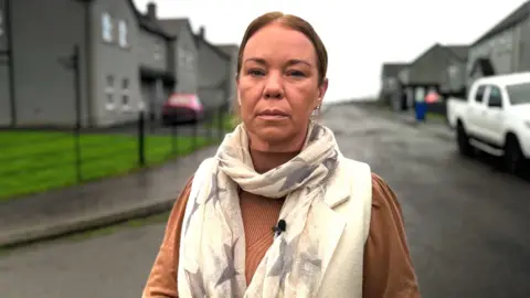 BBC A woman is facing the camera. She has brown hair that is tied up. She's wearing a light brown long-sleeve top and a cream scarf with grey stars. The background is blurred but shows a street of dark grey houses. There is a white truck to the side. 