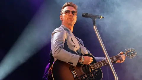Getty Images Ronan Keating performing onstage. He's wearing a suit and holding a guitar. A mic is in front of him. Smoke is in the air and a spotlight is on him. He's wearing glasses.