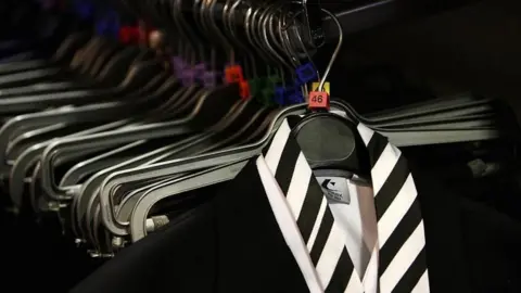 Getty Images A rail with hangers, at the front is a black blazer, white shirt and black and white striped tie
