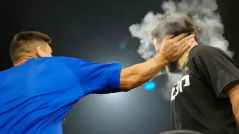 Getty Images A man in a blue t-shirt slapping a man in a black t-shirt with white writing on it. He is slapping him with his right hand. A cloud of chalk dust and a man's hand is obscuring the other man's face.