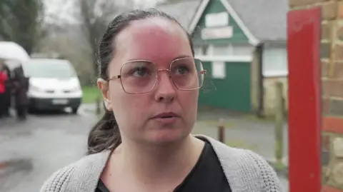 BBC A woman wearing glasses, a black top and a grey cardigan. There is a white van in the background.