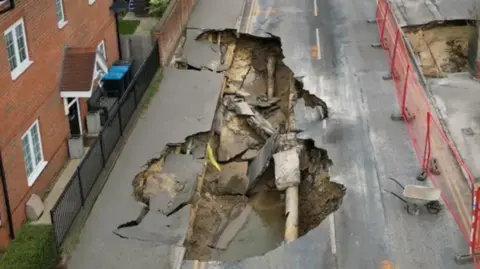 Reuters An aerial picture of a large sinkhole which has opened up beneath one lane of the road. The road surface has collapsed into the hole and a broken pipe and small pool of water are visible. Two sections of the adjoining pavement have also begun to collapse into the hole.