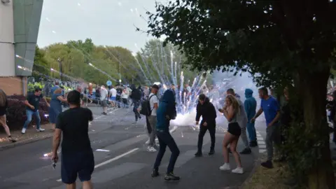 Epitog A group of people stand on a street as a firework explodes in the middle of a road outside a hotel building. Several are turning away amid the explosion.