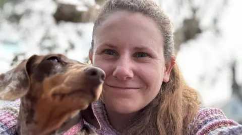 Contributor picture Molly Leonard with a tan and brown Dachshund. Molly is smiling at the camera wearing a pink, purple and yellow knitted jumper, her long brown hair is tied back.