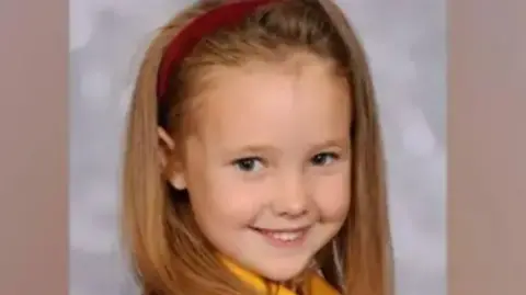 Family handout Elsie Dot Stancombe, wearing a red hairband and her school uniform, smiles as she is photographed.