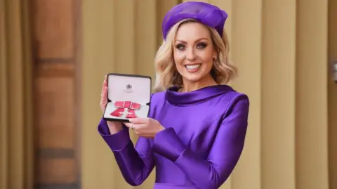PA Media Amy Dowden smiles as she holds her MBE in its presentation box. She is wearing a purple silk dress with a matching purple hat.