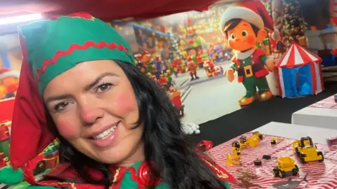 Amanda Walker Selfie of Amanda Walker dressed as an elf. She has long black hair and is wearing a red and green bobble hat. She has red make-up on her cheeks and nose. The room behind her is decorated with a Christmas toy theme.
